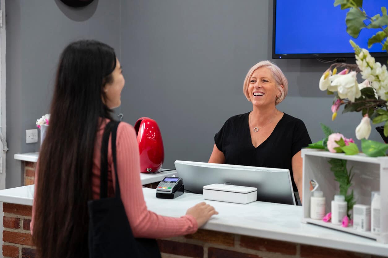 Joyful salon owner at reception desk engaging with a client, epitomising the seamless client commitment and scheduling efficiency made possible by a salon's online booking system, illustrating the article's focus on deposit advantages and salon software.