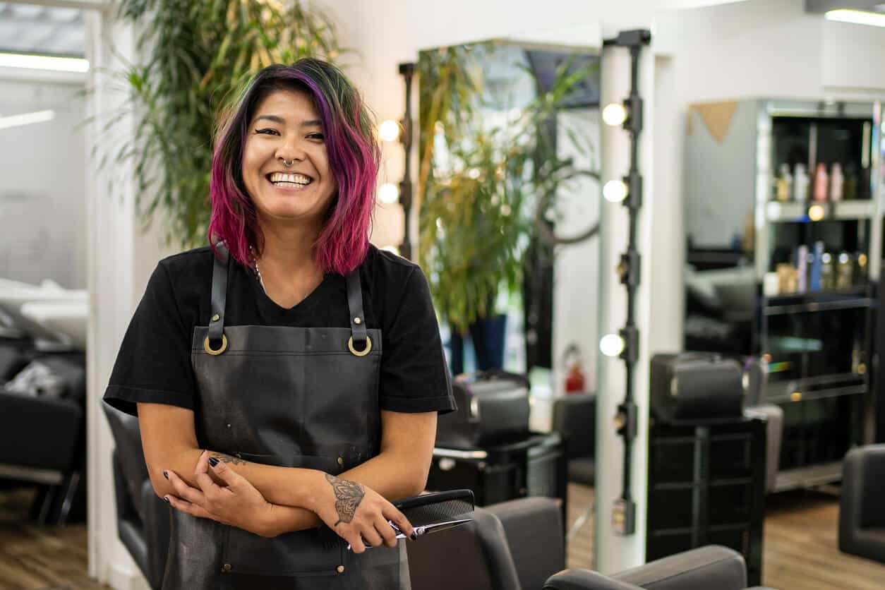 "Cheerful hairdresser with pink hair in a salon, embodying salon online booking success, stands ready in a modern salon setting equipped with the latest beauty salon software features, embodying efficient salon scheduling and booking system."