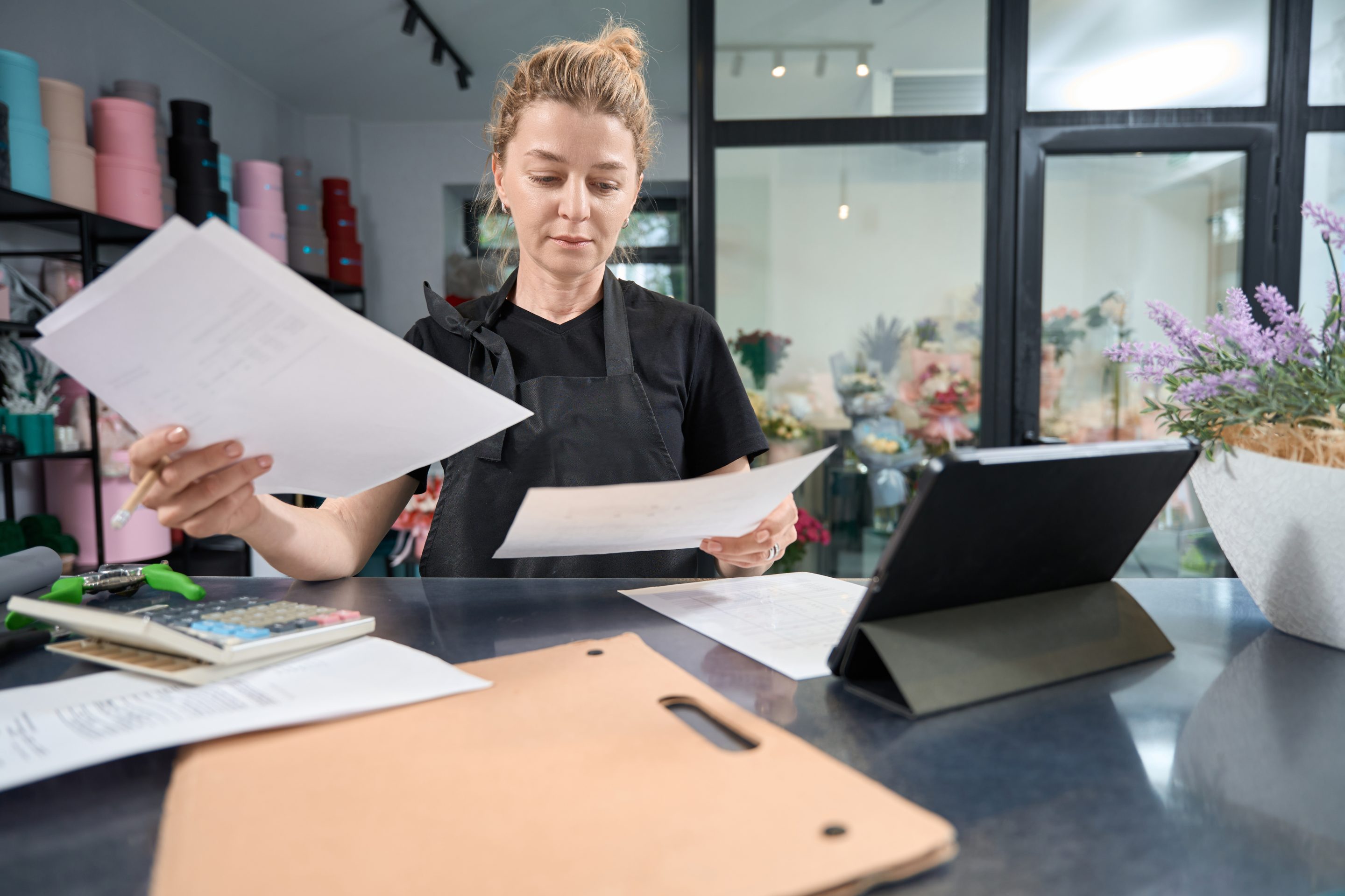 Focused flower shop owner sitting at counter looking through paperwork A salon owner surrounded by paperwork and seemingly overwhelmed, highlighting the time-consuming administrative tasks. Discover how salon management software can streamline these processes, freeing up valuable time to focus on clients and grow your business.