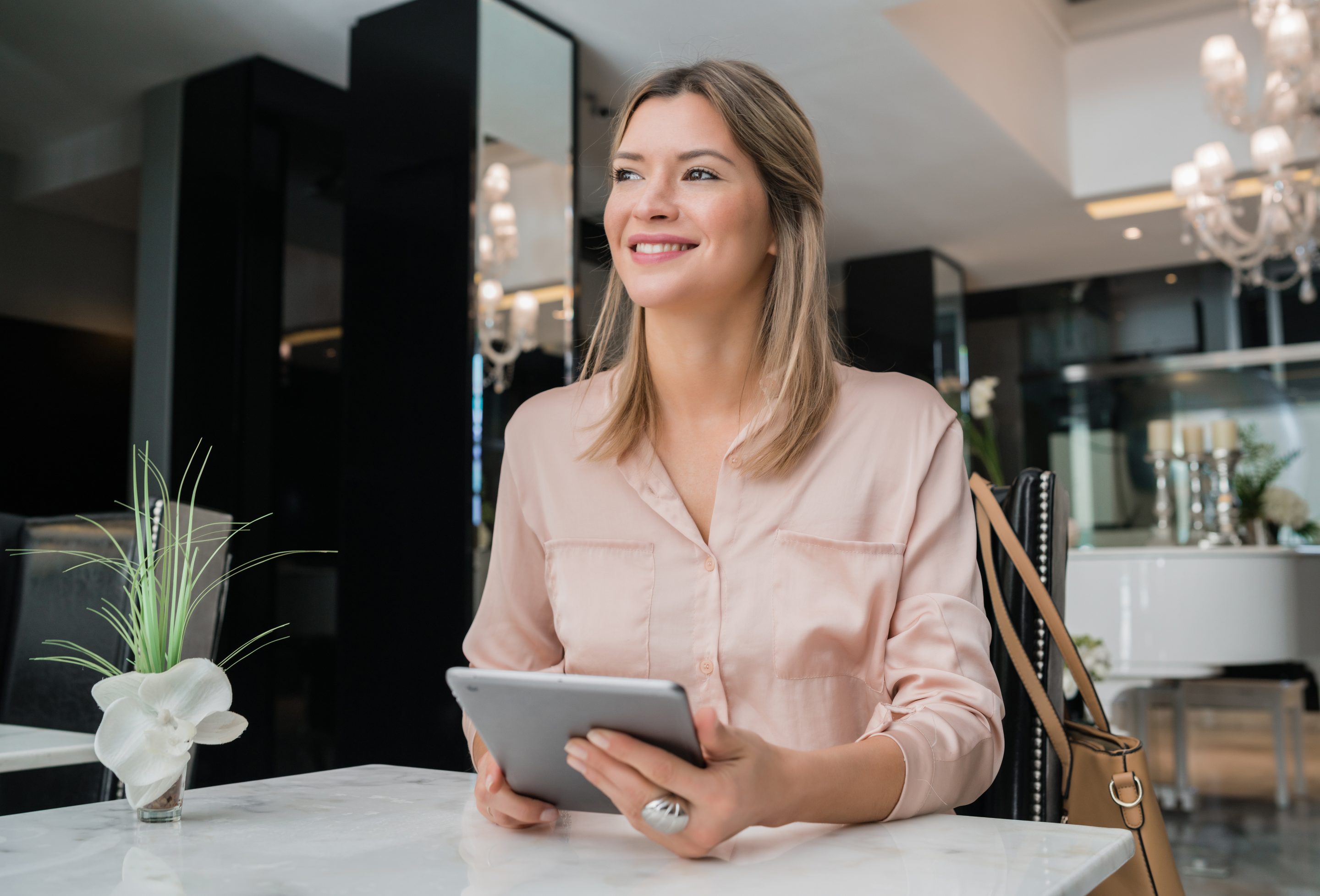 businesswoman working on her digital tablet. A smiling woman in a light pink blouse sits at a table in a modern salon, holding a tablet, symbolizing confidence and efficiency in managing salon operations.